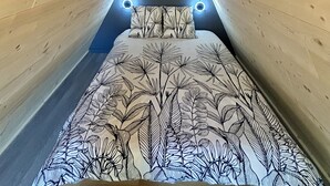 Een slaapkamer met een schuine plafond, een bed met een sprei met bladmotief, twee wandlampen en een raam aan het uiteinde van de kamer.