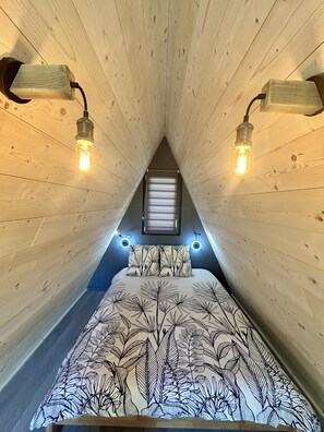 A bedroom with a sloped ceiling, a bed with a leaf-patterned bedspread, two wall-mounted reading lights, and a window at the end of the room. - A-Frame House with Mountain View, Private Terrace, and Wi-Fi (Touët-sur-Var)