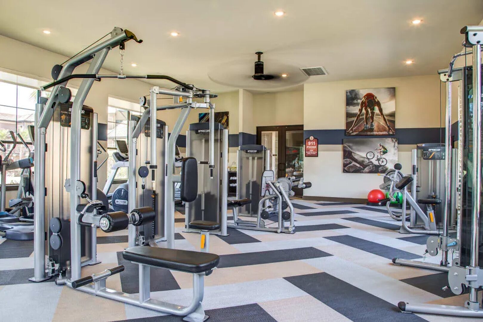 A gym with various exercise machines, a bench, and a wall-mounted sign indicating an emergency phone.