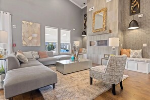 Ein modernes Wohnzimmer mit einem grauen Ecksofa, einem gemusterten Sessel und einem Couchtisch. An den Wänden befinden sich abstrakte Kunstwerke und ein dekorativer Spiegel.
