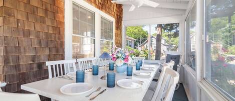 Un espace extérieur abrité, aménagé pour les repas, avec une table blanche dressée pour six personnes, entourée de chaises blanches, d’un ventilateur de plafond et de grandes fenêtres.