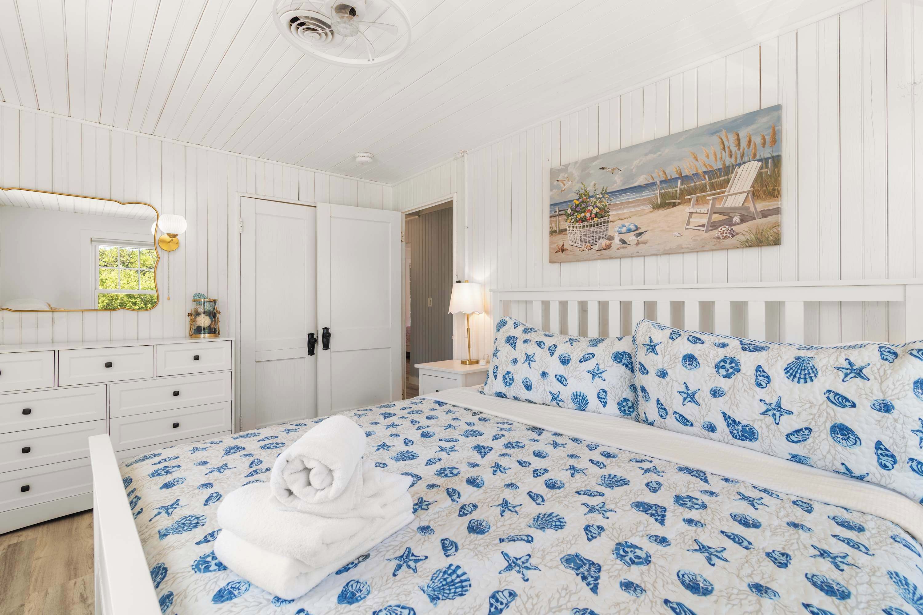 A bedroom with a bed, white walls, a painting of a beach scene, and a nightstand with a lamp.