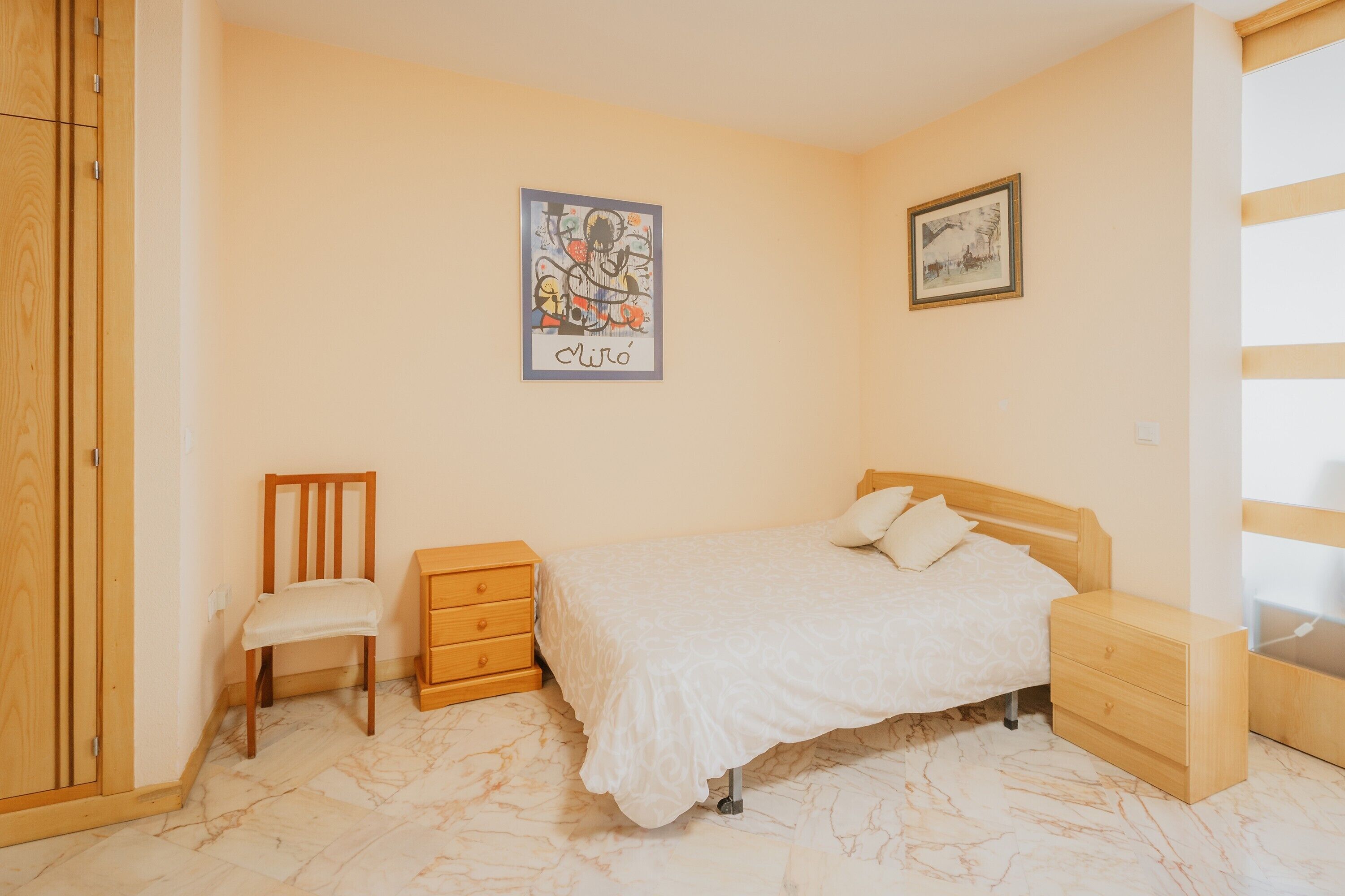 Ein Schlafzimmer mit einem Bett, einem Sessel, einem Nachttisch, einem Kleiderschrank und zwei Bildern an der Wand.
