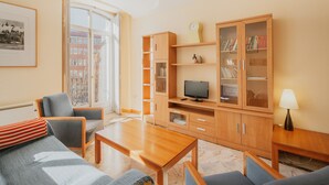 Een woonkamer met een bank, stoelen, een houten tv-meubel en een raam met uitzicht op gebouwen.