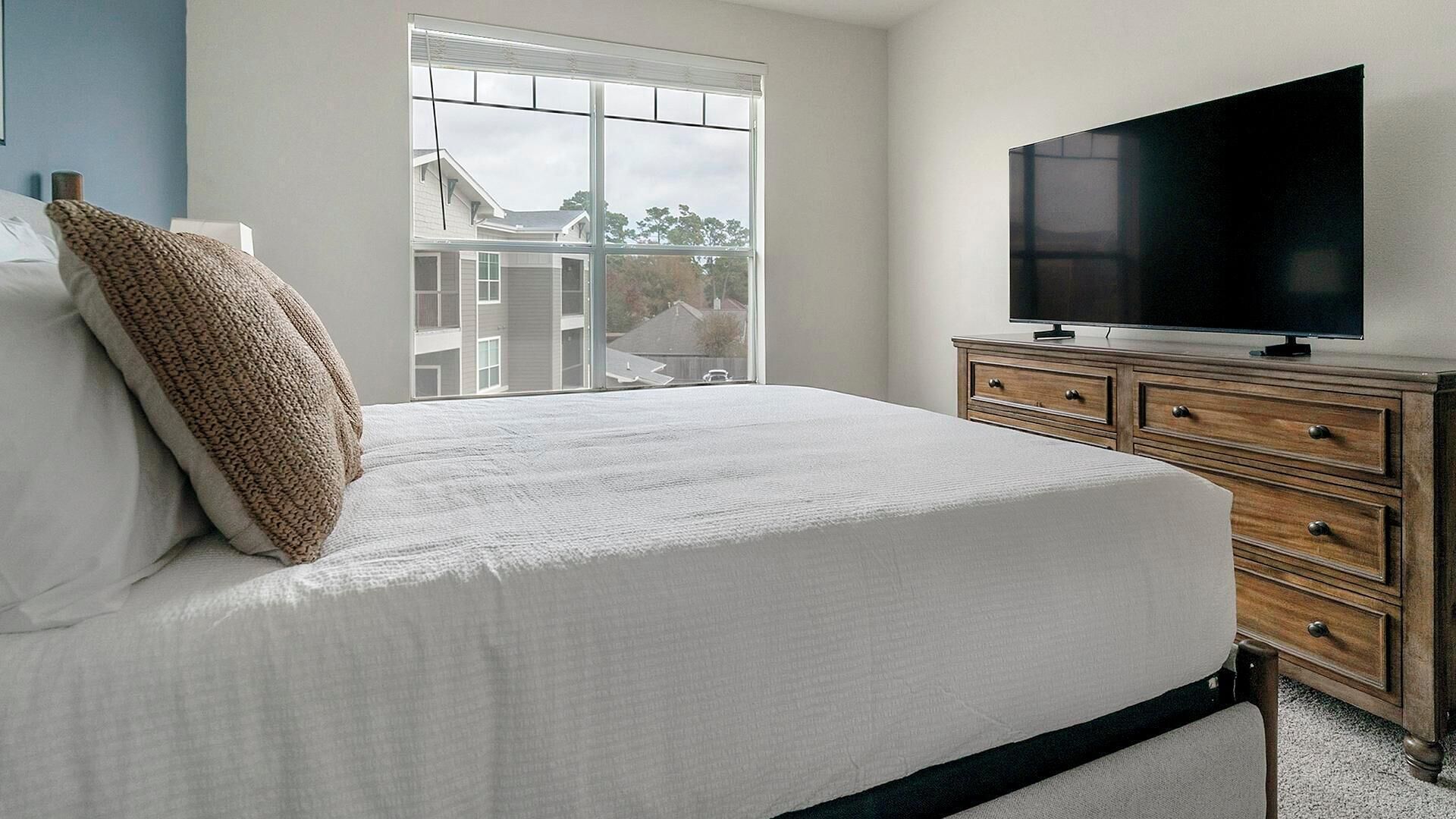 Une chambre à coucher comprenant un lit, une commode, un téléviseur et une fenêtre donnant sur des immeubles.