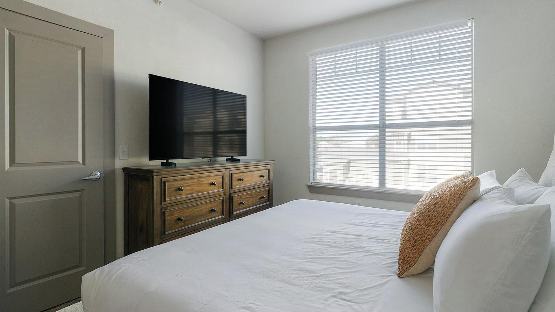 Een slaapkamer met een bed, een dressoir, een televisie en een raam met jaloezieën.