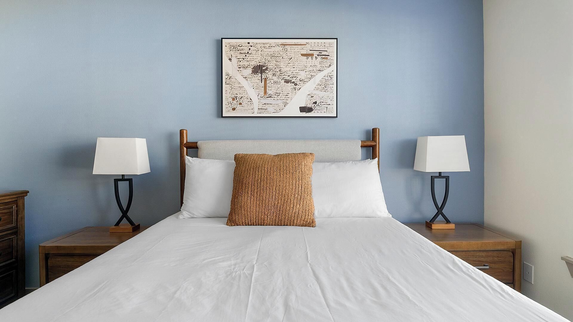 Een net opgemaakt bed met witte lakens, een geweven sierkussen en twee bedlampjes op houten nachtkastjes. Daarboven hangt een lijst met abstracte kunstwerken.