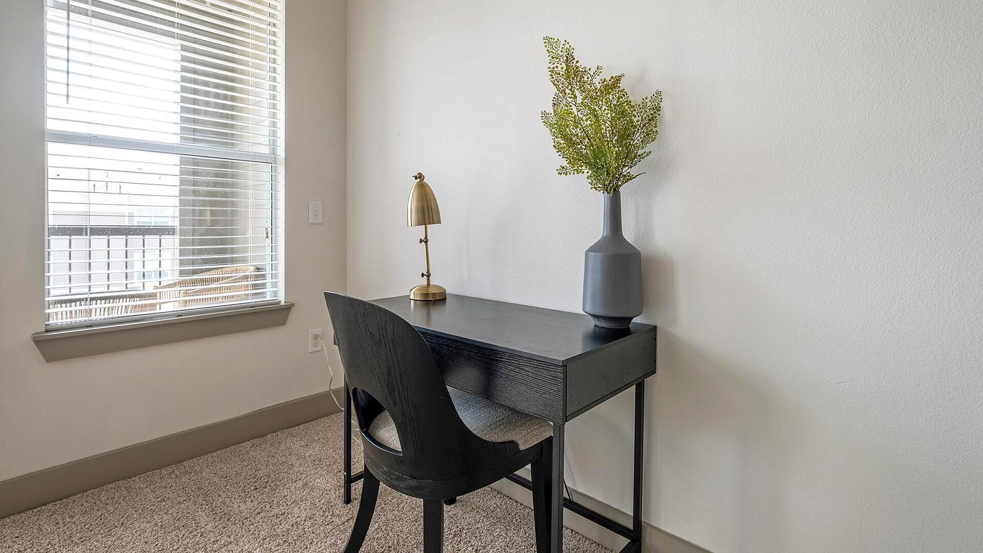 A small office space with a desk, chair, lamp, and a vase with a plant.