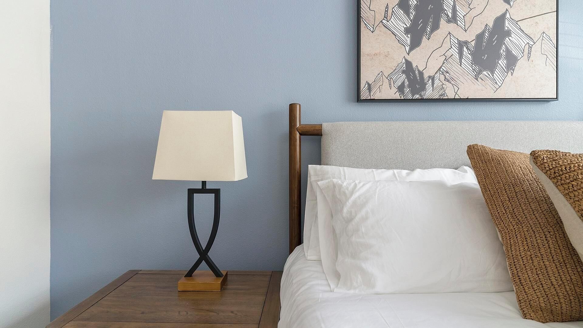 Een net opgemaakt bed met witte en bruine kussens, een houten nachtkastje met een moderne lamp, en een blauwe wand met een ingelijst abstract kunstwerk.