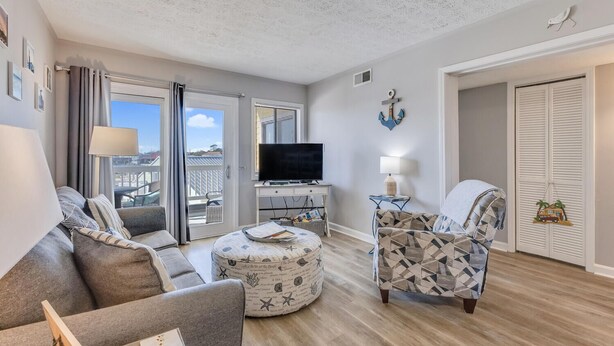 Ein Wohnzimmer mit Sofa, Hocker und einem gemusterten Sessel, einem Fernseher und einem Balkon mit Blick.