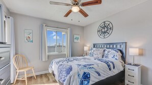 Ein Schlafzimmer mit einem Bett, einem Holzsessel, einem Nachttisch, einem Deckenventilator, einem Fenster mit Vorhängen und Wandbildern.