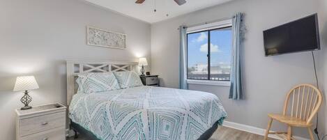 Un dormitorio con cama, mesitas de noche, ventilador de techo, televisor y ventana con cortinas.