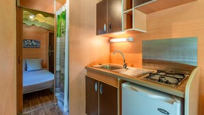 Een kleine keuken met spoelbak, kookplaat en kastjes, naast een bed in een eenvoudige kamer.