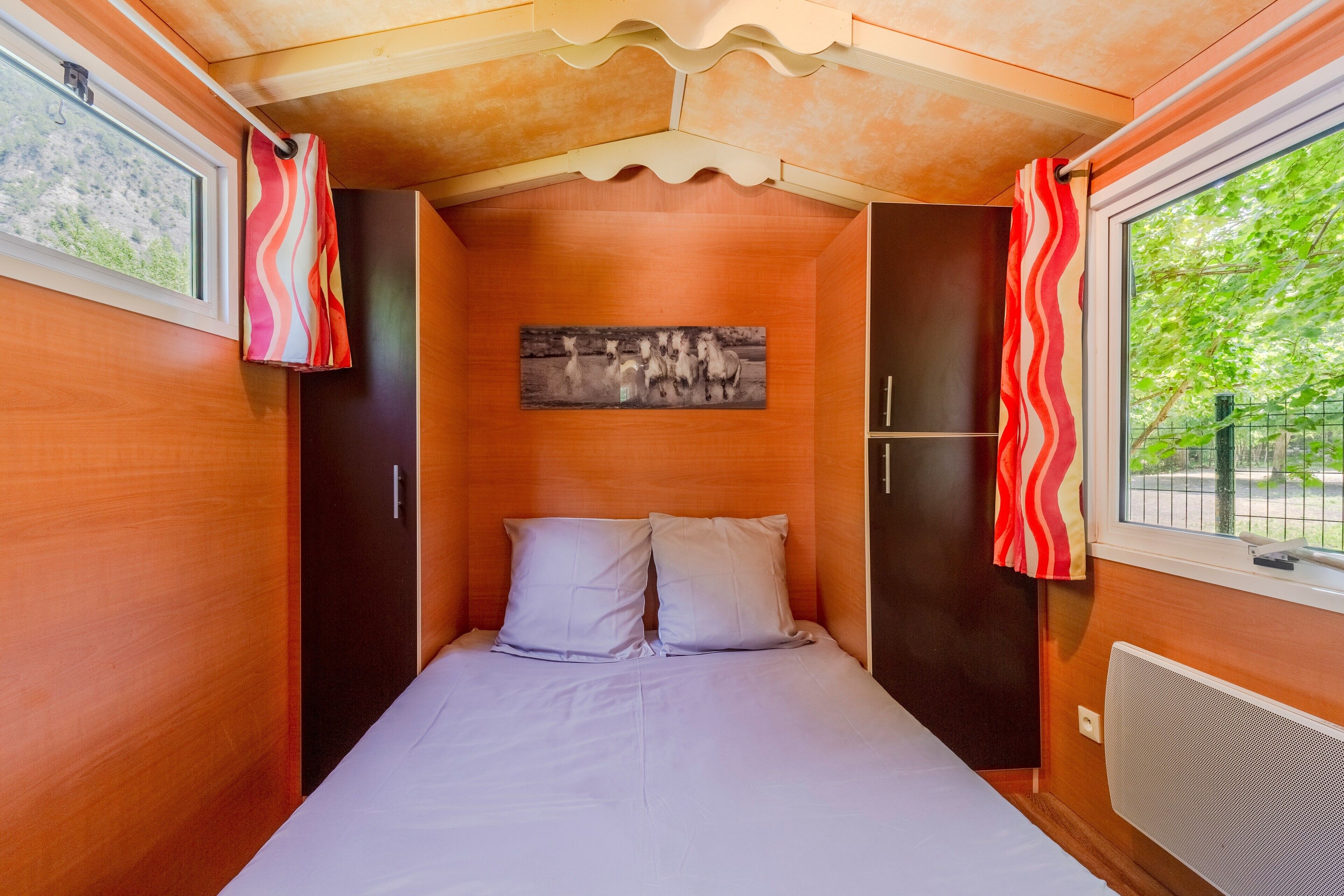 A narrow, single-bed room with a wooden ceiling, a window with a view of greenery, and a wall-mounted artwork.