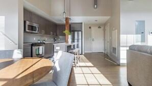A modern kitchen with stainless steel appliances, a dining area with a wooden table and grey upholstered chairs, and a large window allowing natural light. - Landing | Stunning 1BD, Gym, Game Room (New Haven)