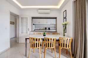 A dining area with a white table, wooden chairs, a vase with flowers, and a kitchen in the background. - Apartment 'Golf y Mar - T2' with Shared Pool, Wi-Fi and Air Conditioning (Vilamoura)