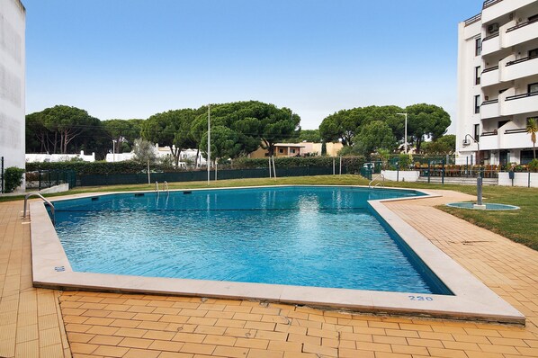 A rectangular swimming pool with a tiled deck, surrounded by a fence, and trees in the background. - Apartment 'Golf y Mar - T2' with Shared Pool, Wi-Fi and Air Conditioning (Vilamoura)