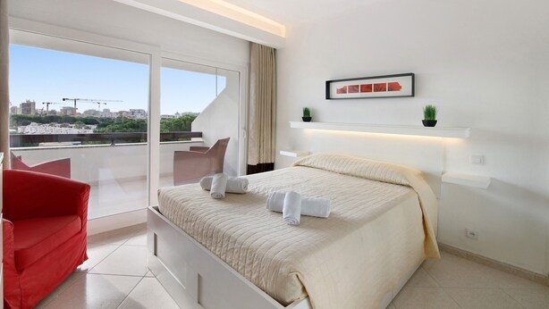 Ein modernes Hotelzimmer mit einem großen Bett, einem roten Sessel und Blick auf die Stadt vom Balkon.