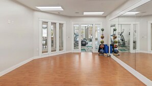 Amplia habitación con piso de madera, paredes blancas y un espejo grande. Se ven pelotas de ejercicio y una caminadora a través de puertas de vidrio.