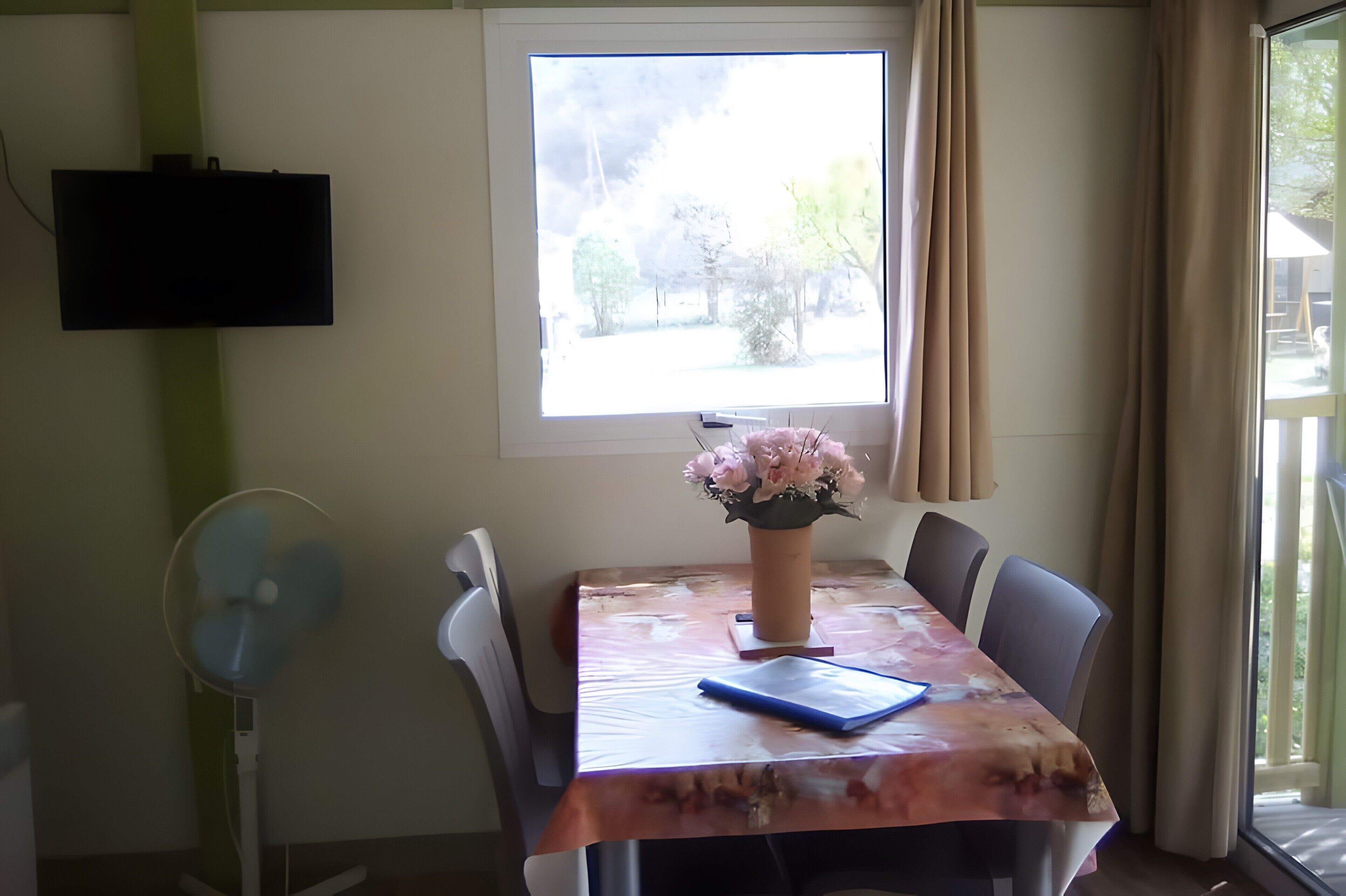 Uma sala de jantar com mesa, cadeiras, um vaso de flores, televisão na parede e uma janela com cortinas.