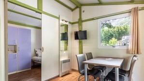 A room with a wooden floor, a table with chairs, a TV mounted on the wall, a window with curtains, and a bed with a pillow. - Holiday Home 'Chalet 1 À 6 Personnes Pmr Access' with Private Terrace and Wi-Fi (Touët-sur-Var)