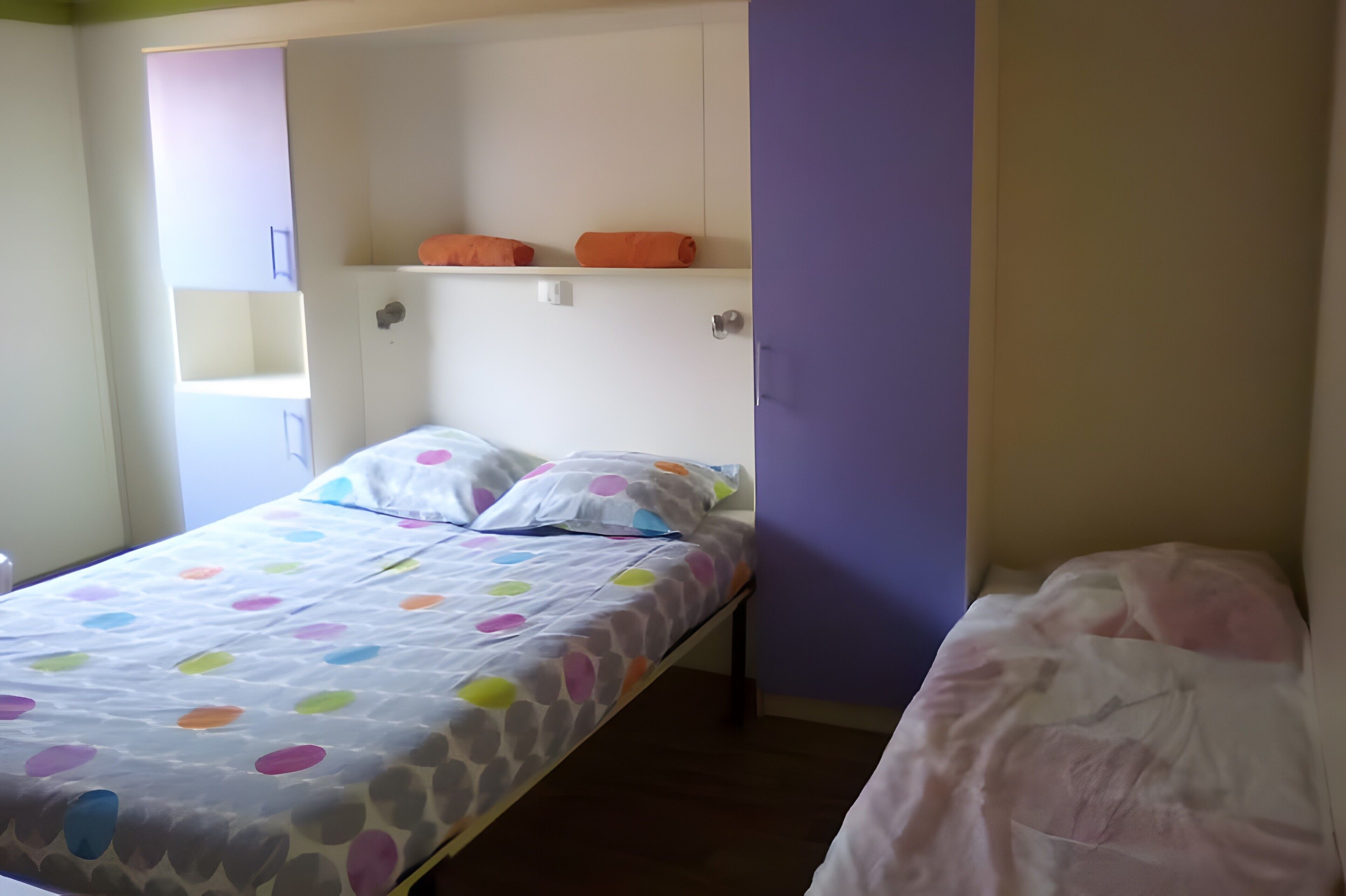 Een eenpersoonsbed met een stippenbedekking, een klein plankje met oranje handdoeken, en een paarse muur.