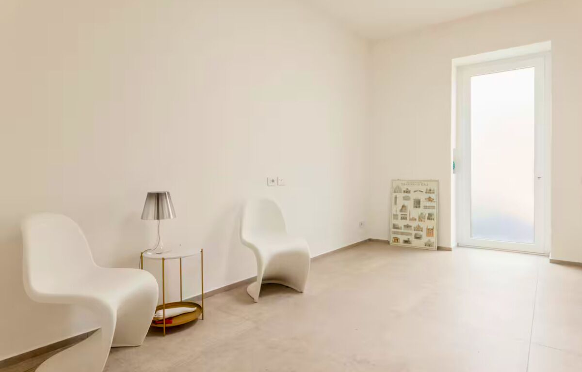 A minimalist room with two white chairs, a small round table with a lamp, and a door leading to a balcony.