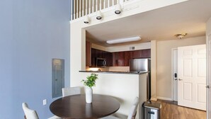 Un coin repas avec une table ronde, quatre chaises, un vase avec une plante, et une cuisine dotée de meubles en bois foncé et d’appareils électroménagers en acier inoxydable.
