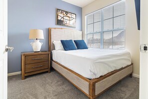 Ein Schlafzimmer mit Bett, Nachttisch, Lampe, Wandbild und einem Fenster mit Jalousien.