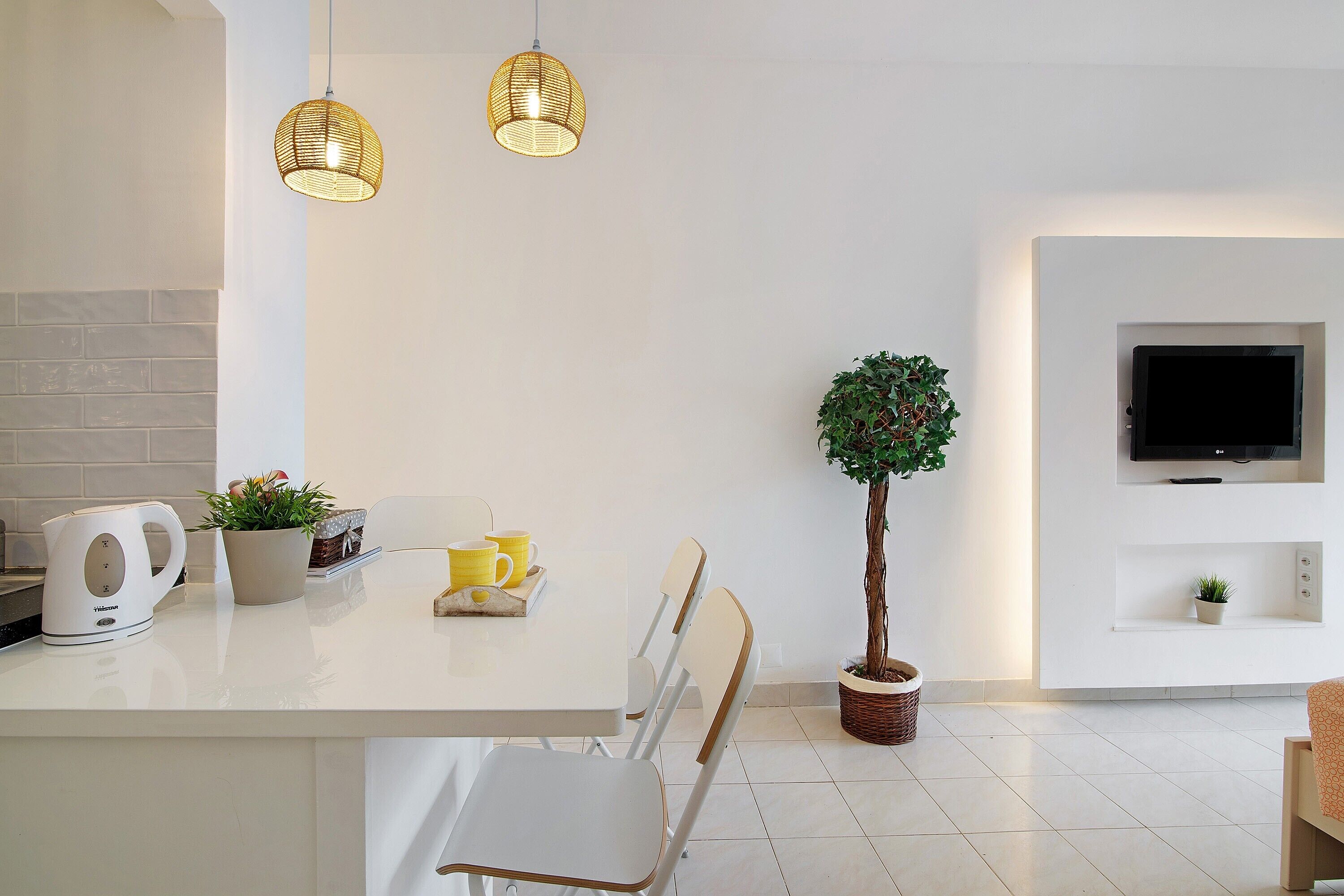 Une cuisine moderne avec un plan de travail blanc, deux luminaires suspendus en osier, une plante en pot, une bouilloire et un téléviseur fixé au mur, qui est également blanc.