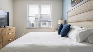 A bedroom with a bed, a TV on a wooden dresser, a window with blinds, and a lamp on a bedside table.