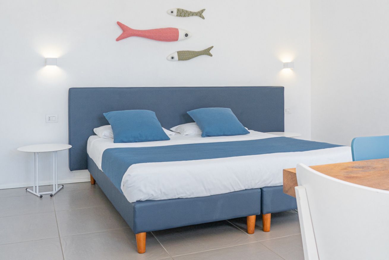 A bed with a blue headboard, two blue pillows, and a white bedspread. There are two wall-mounted lights and two fish-shaped wall decorations.