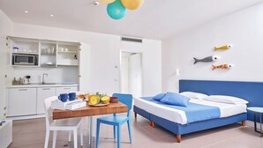 Una habitación de hotel moderna con una cama azul, paredes blancas, una mesa de comedor de madera con sillas, una zona de cocina con armarios blancos y un techo con lámparas esféricas.