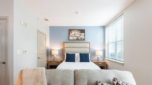 Une chambre d’hôtel comprenant un lit, deux tables de chevet avec des lampes, une grande fenêtre avec des stores, une porte et une œuvre d’art accrochée au mur.