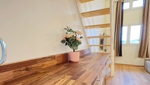 Una habitación amplia con suelo de madera, una escalera con vigas de madera, una planta en maceta y una ventana con cortinas.