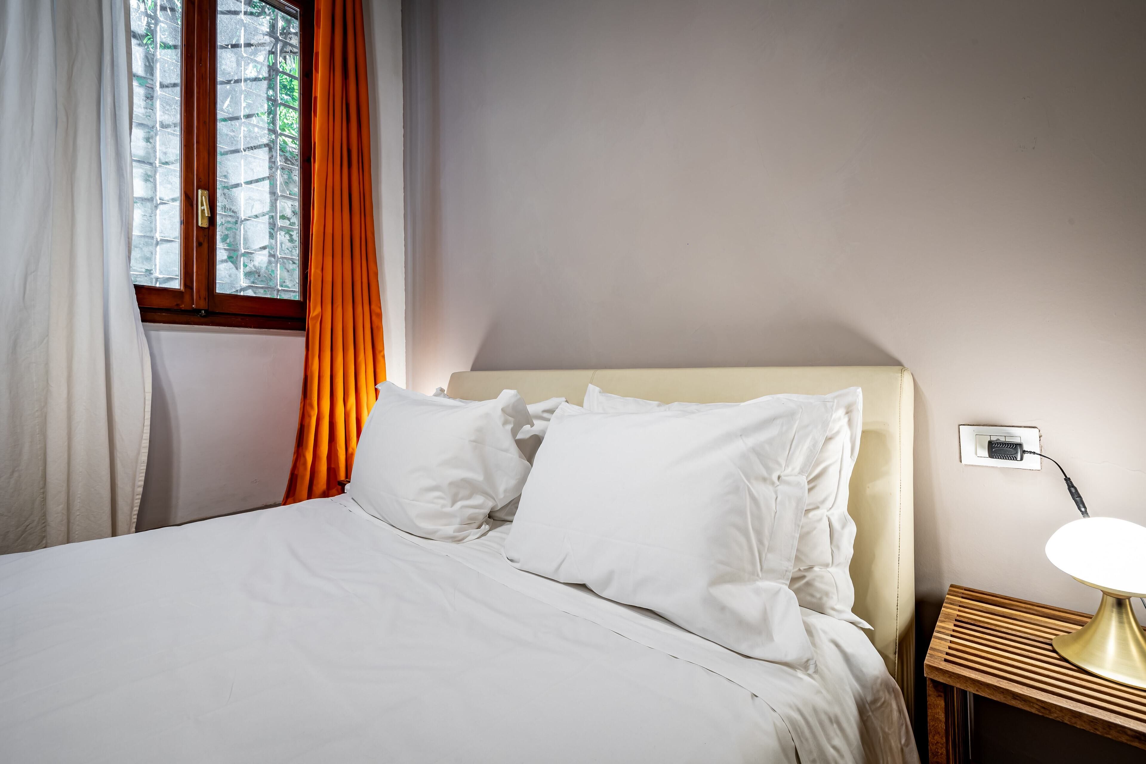 A neatly made bed with white linens and pillows, a bedside table with a lamp, and a window with curtains.