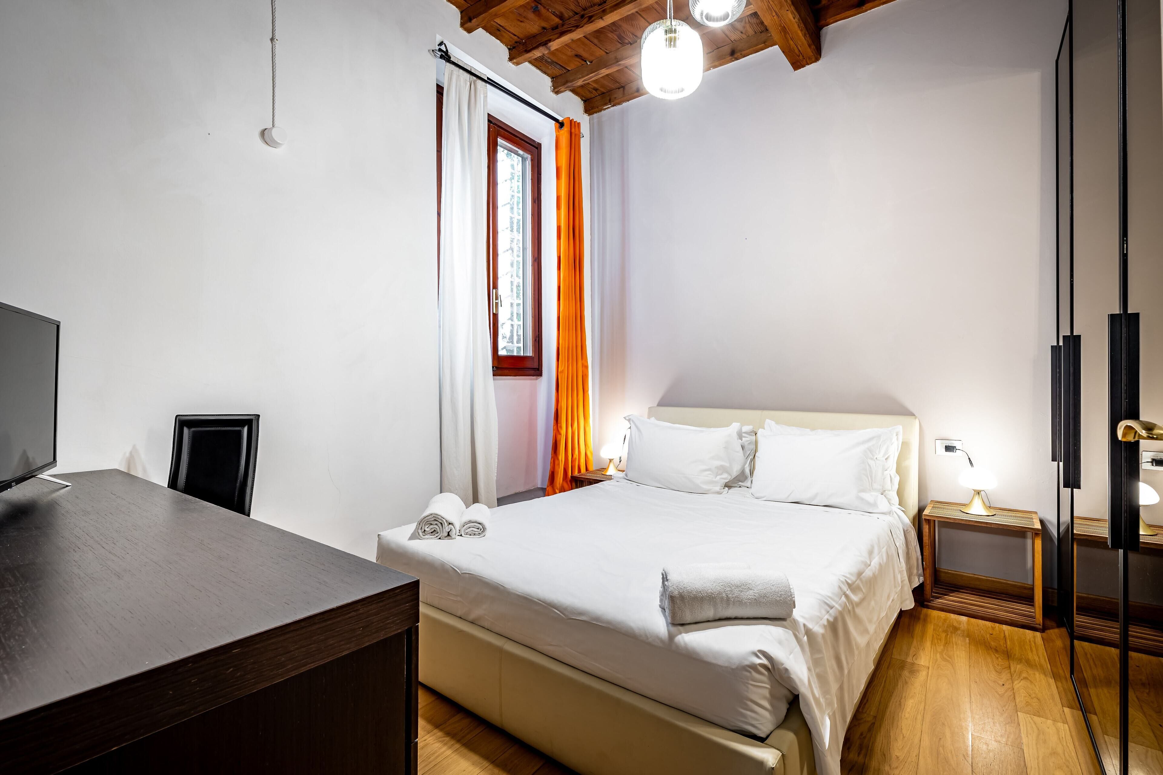 A bedroom with a bed, a desk, a chair, a window with curtains, and a wooden ceiling.