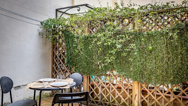An outdoor patio with a round table set for two, surrounded by black chairs, and a wooden lattice fence partially covered with green foliage. - YiD Lungarno Cellini retreat (Florence)