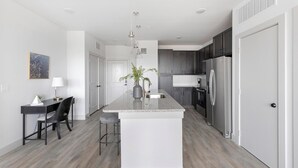 A modern kitchen with a central island, dark cabinets, stainless steel appliances, and a dining area with a table and chairs. - Landing | Stunning 2BD in Corinth (Denton)