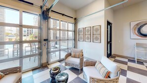 A spacious room with a large sliding glass door leading to an outdoor patio area. There are two wicker chairs and a white sofa with striped pillows. The room has a checkered floor and a wall-mounted shelf. - Landing | Stunning 1BD in Corinth (Denton)