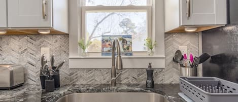 A kitchen with a herringbone tile backsplash, a stainless steel sink, and a window with a view of a building and trees.