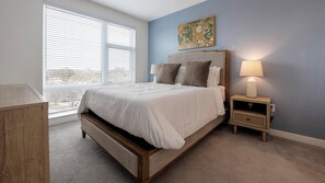 Un dormitorio con una cama grande, una mesita de noche con lámpara, cabecera de madera y una ventana con persianas.