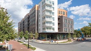 A modern multi-story building with a glass facade, a prominent entrance, and a parking area with a blue utility vehicle. - Landing | Amazing 2BD, Clubhouse, Courtyard (Alexandria)