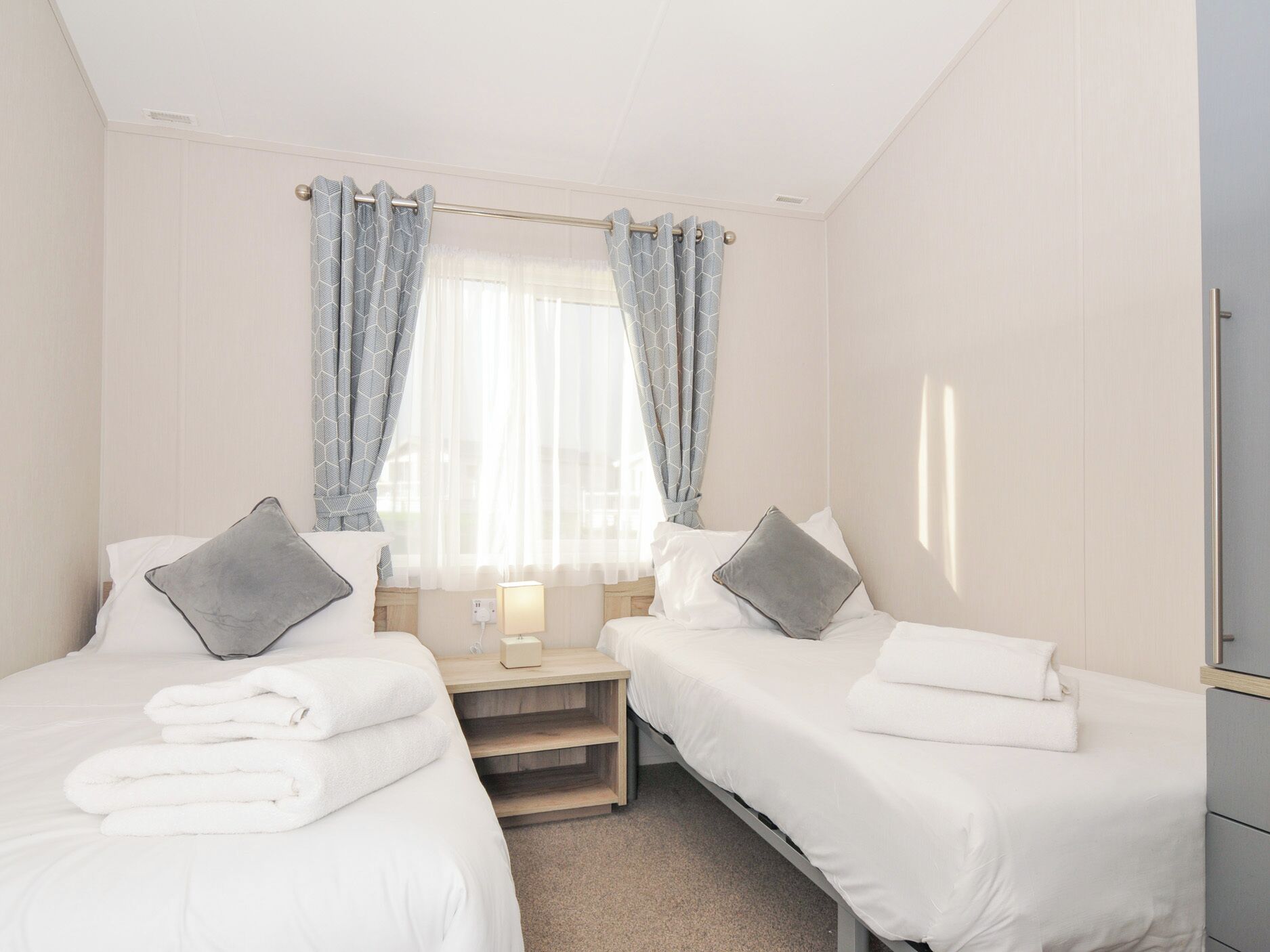 Habitación de hotel con dos camas individuales, una mesita de noche con lámpara y una ventana con cortinas.