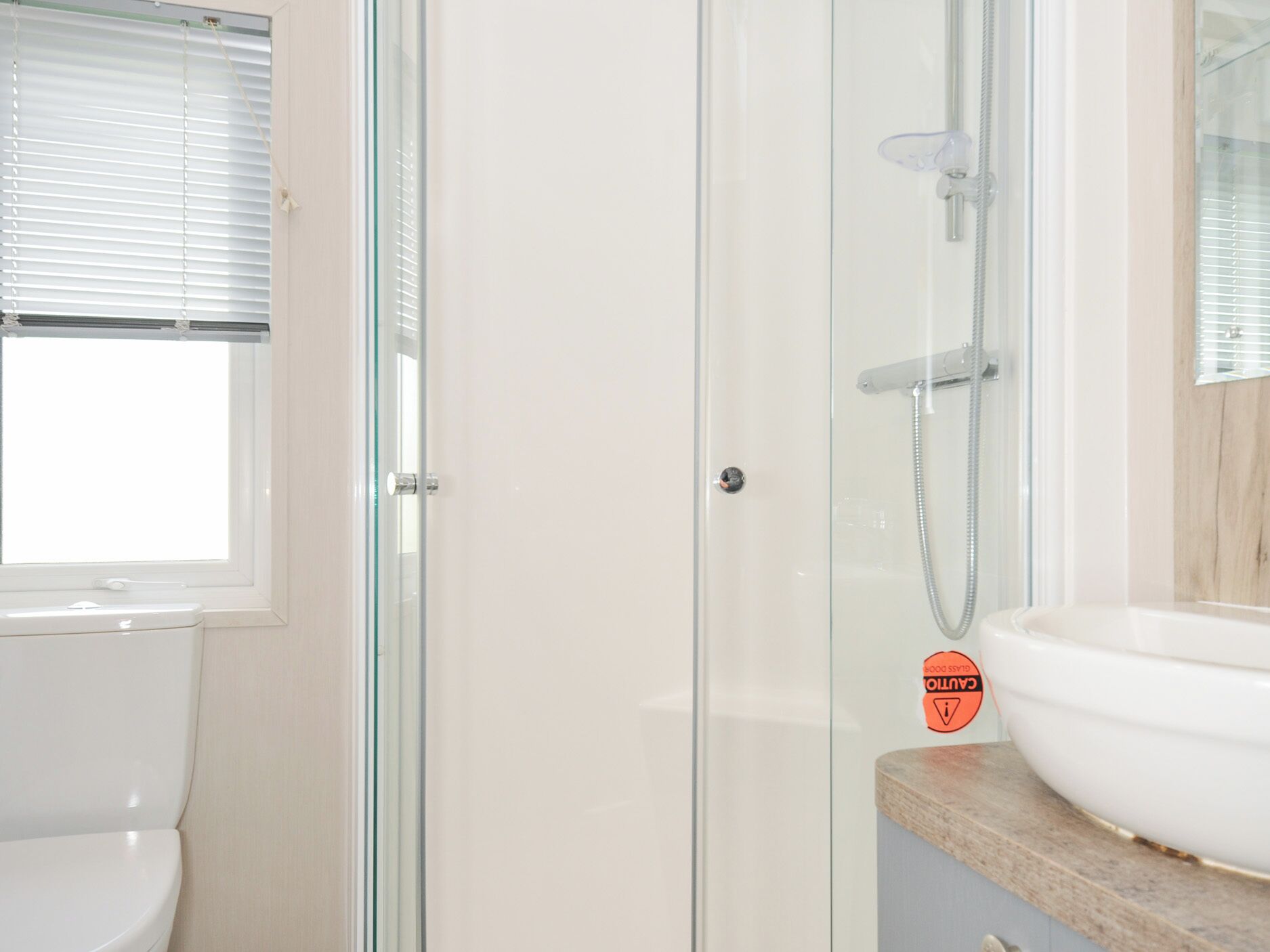 A bathroom with a glass shower enclosure, a white sink, and a window with blinds.