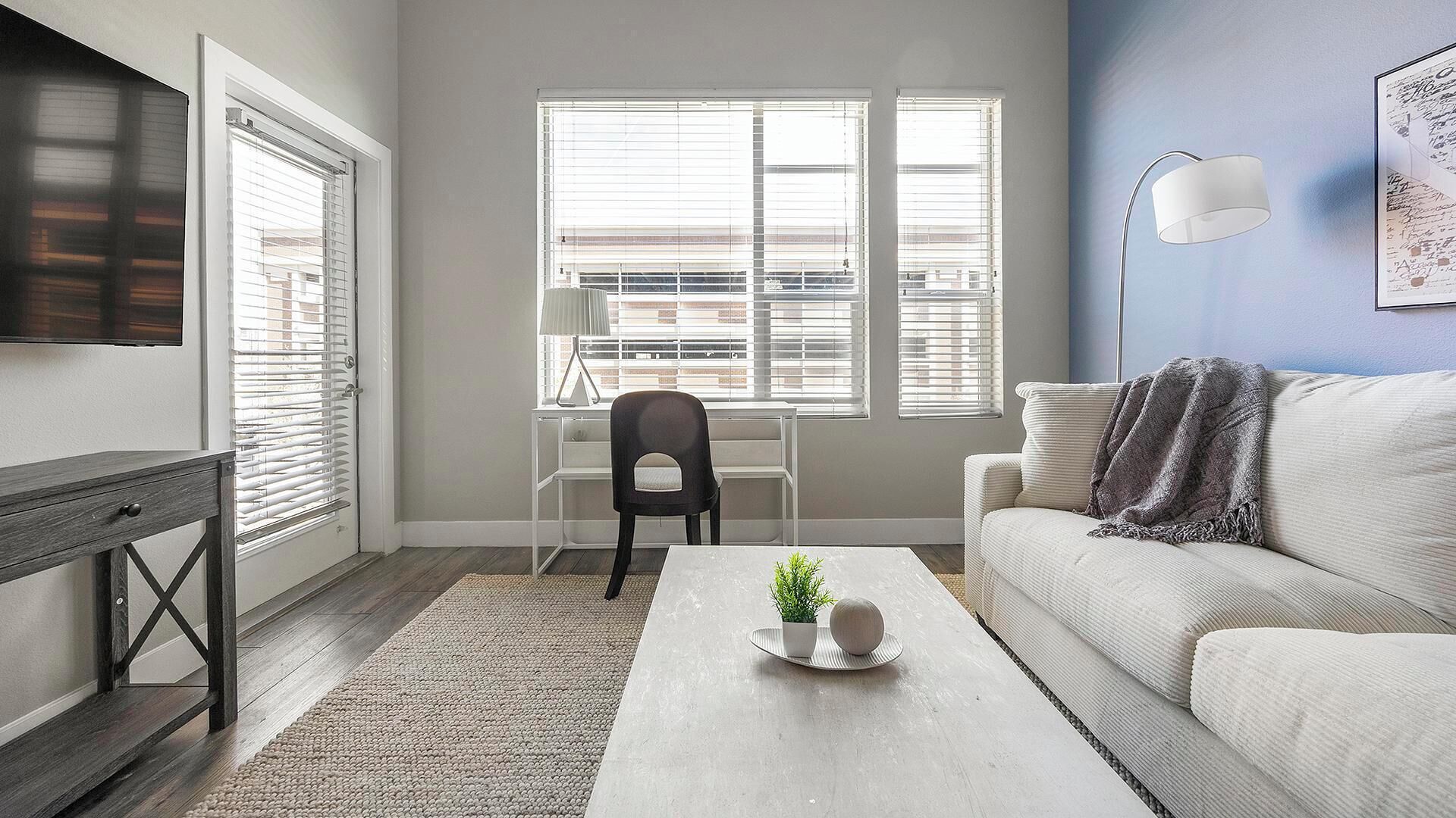 A modern living room with a white sofa, a coffee table with a plant, and a chair. There are large windows with blinds, a TV mounted on the wall, and a floor lamp.