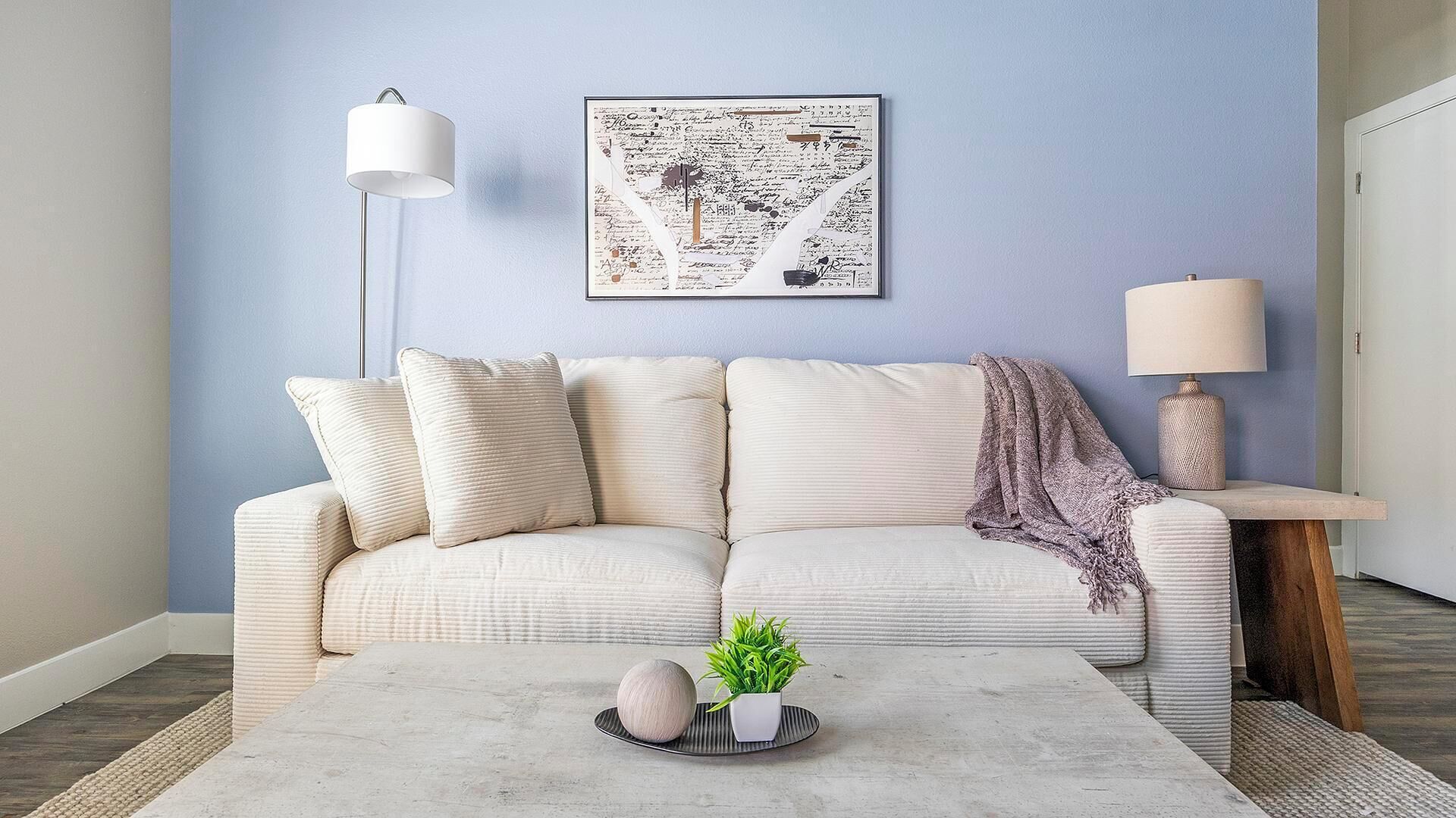 A living room with a light blue wall, a white sofa with cushions, a coffee table with decorative items, a floor lamp, and a side table with a lamp.