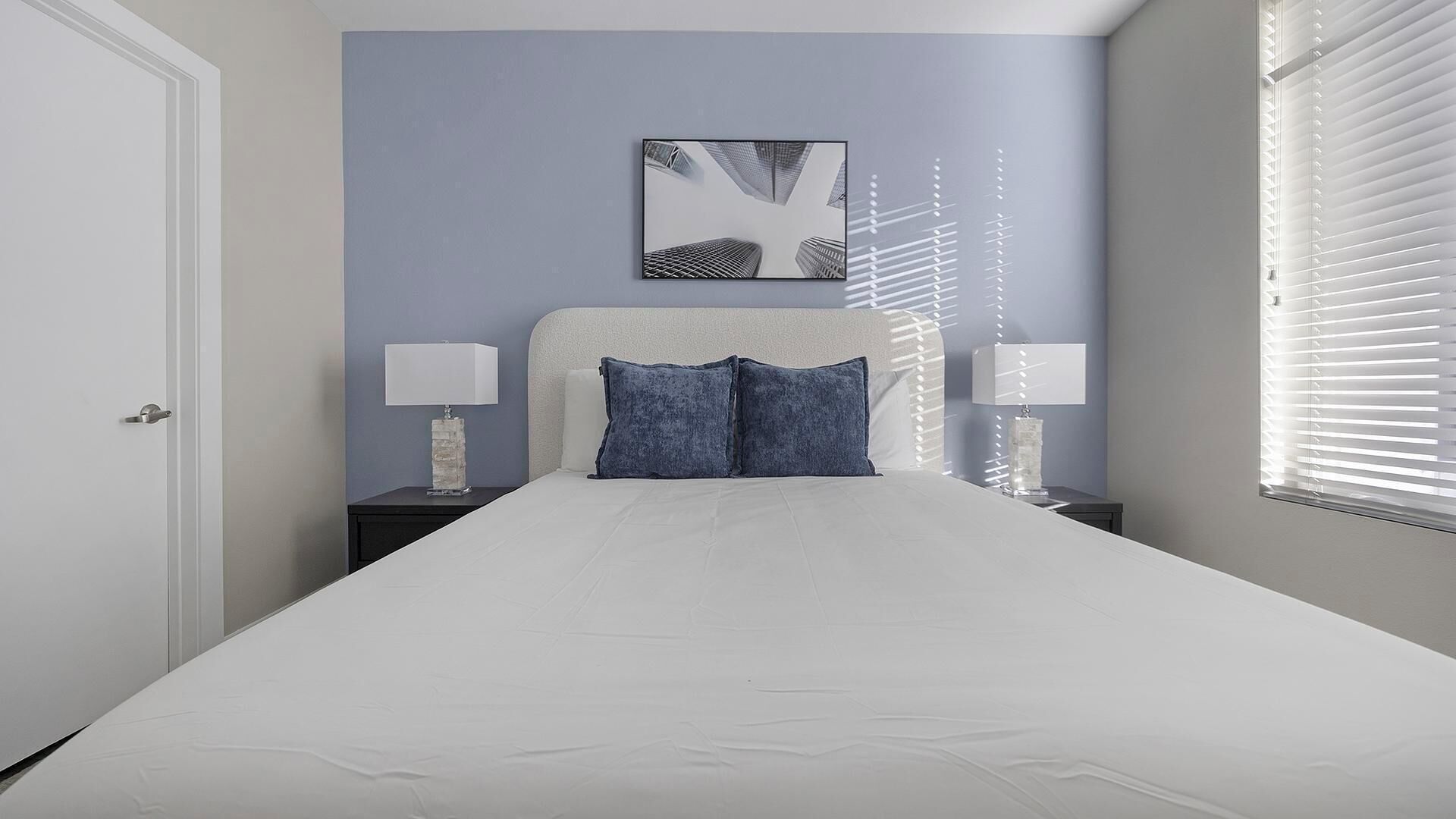 A bedroom with a bed, two nightstands, a headboard, a wall-mounted artwork, and a window with blinds.