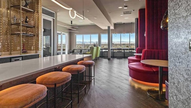 A modern bar area with a long counter, multiple stools, and a view of the cityscape through large windows. - Landing | Modern 2BD, Gym, Clubhouse (Denver)