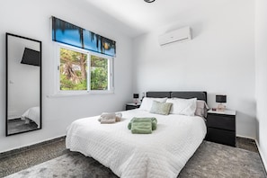 Ein Schlafzimmer mit einem Bett, zwei Nachttischen, einem Wandgerät zur Klimatisierung und einem Spiegel, durch ein Fenster blickt man auf Grünflächen.